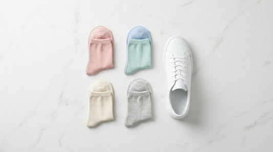 Five pairs of women's ankle socks in soft pastels and neutrals laid out in a clean grid on white marble beside a single white sneaker