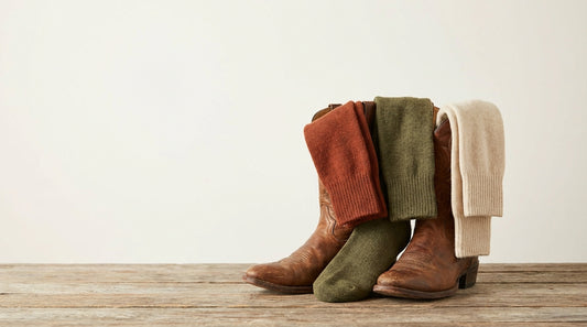 Premium over-the-calf boot socks in earth tones draped over a pair of classic western cowboy boots on a rustic surface