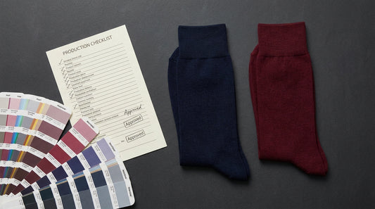 Editorial flat-lay photograph of premium custom dress socks in navy and burgundy arranged on a matte charcoal surface beside a production checklist and Pantone TCX swatch book, studio lighting, manufacturer's quality-control scene