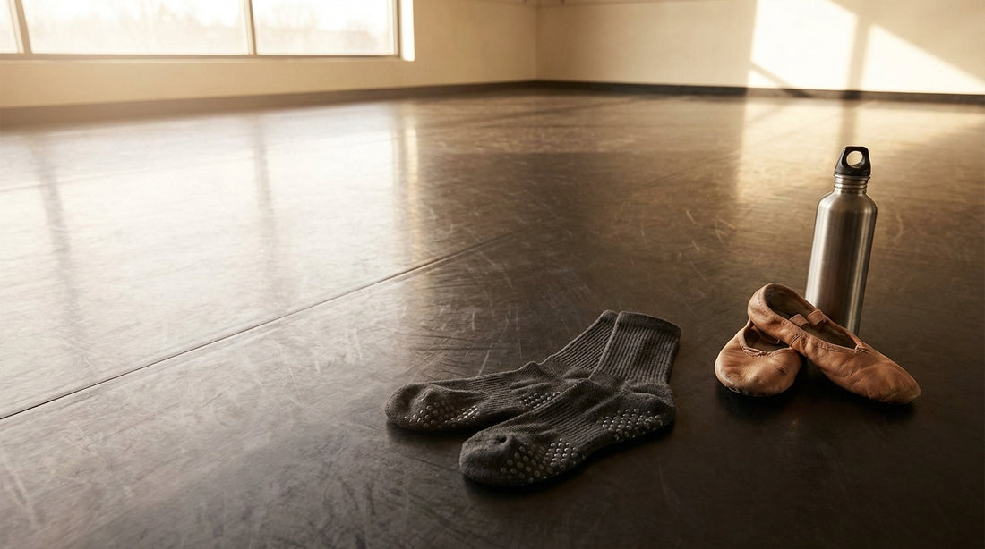 Grip socks with silicone dot patterns laid on a polished dance studio marley floor next to ballet shoes and a water bottle