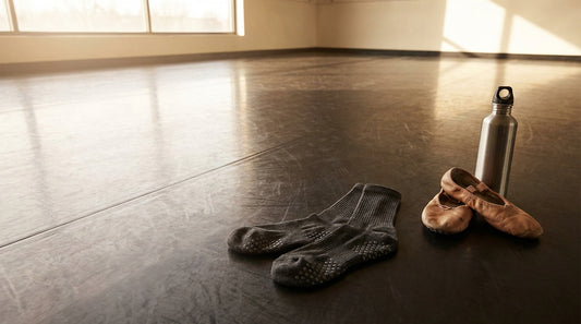 Grip socks with silicone dot patterns laid on a polished dance studio marley floor next to ballet shoes and a water bottle