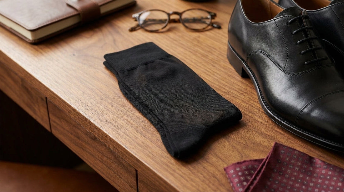 Folded fine-denier black sheer dress socks on a polished walnut desk beside a black leather dress shoe and a burgundy silk pocket square