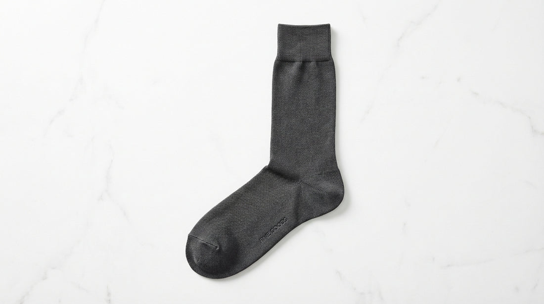 Premium dress sock laid flat showing six quality details: fine-gauge knit texture, reinforced heel, seamless toe, ribbed cuff, fabric sheen, and branded sole