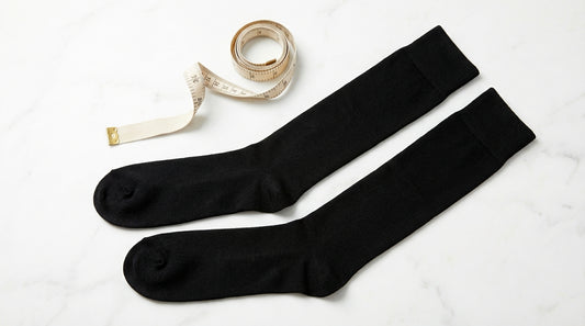 A pair of premium black over-the-calf dress socks laid flat on a white marble surface with a fabric measuring tape curled beside them