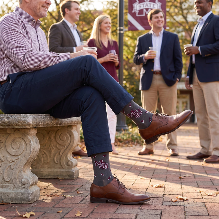 MSU Gray Cowbell Dress socks worn seated, showing scattered maroon cowbells and the interlocking MSU logo on charcoal
