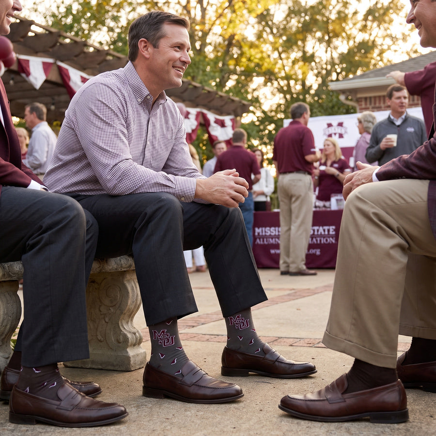 MSU Gray Cowbell Dress socks worn seated, second angle, showing scattered maroon cowbells and the interlocking MSU logo on charcoal