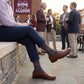 MSU Gray Cowbell Dress socks worn seated, third angle, showing scattered maroon cowbells and the interlocking MSU logo on charcoal