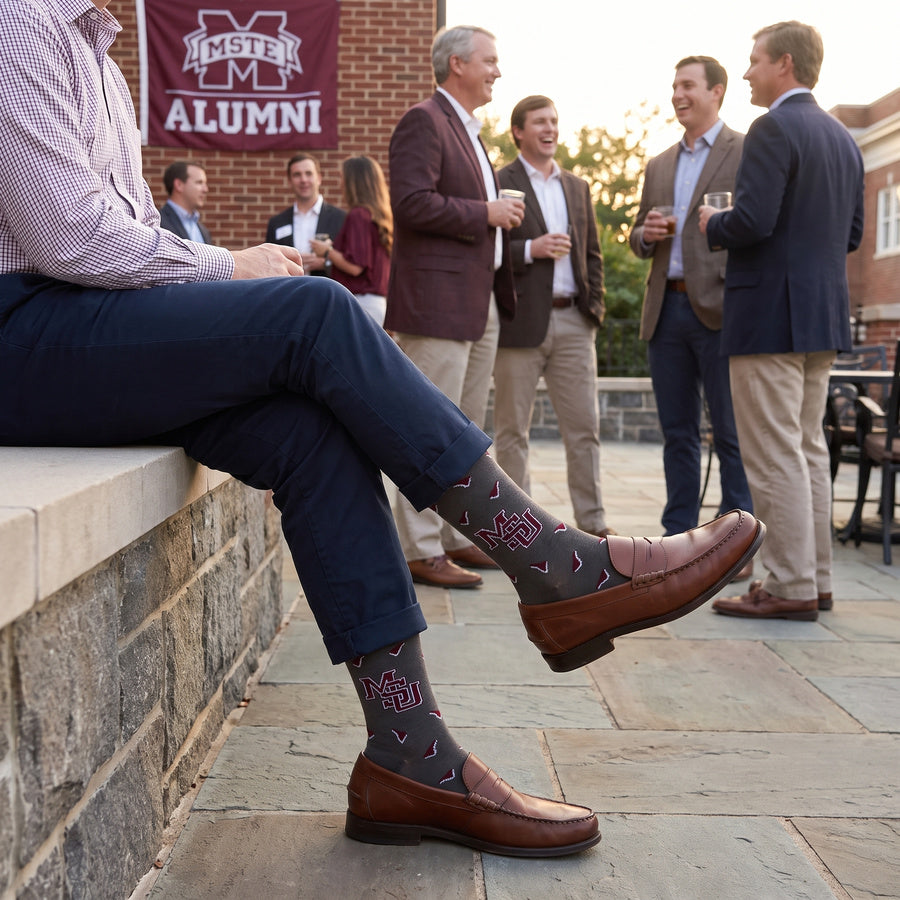 MSU Gray Cowbell Dress socks worn seated, third angle, showing scattered maroon cowbells and the interlocking MSU logo on charcoal