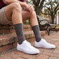 MSU Gray Cowbell Dress socks worn seated on campus, showing scattered maroon cowbells and the interlocking MSU logo on charcoal