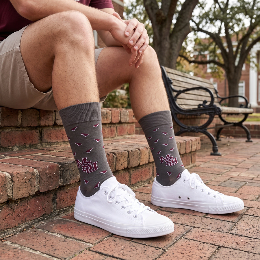 MSU Gray Cowbell Dress socks worn seated on campus, showing scattered maroon cowbells and the interlocking MSU logo on charcoal