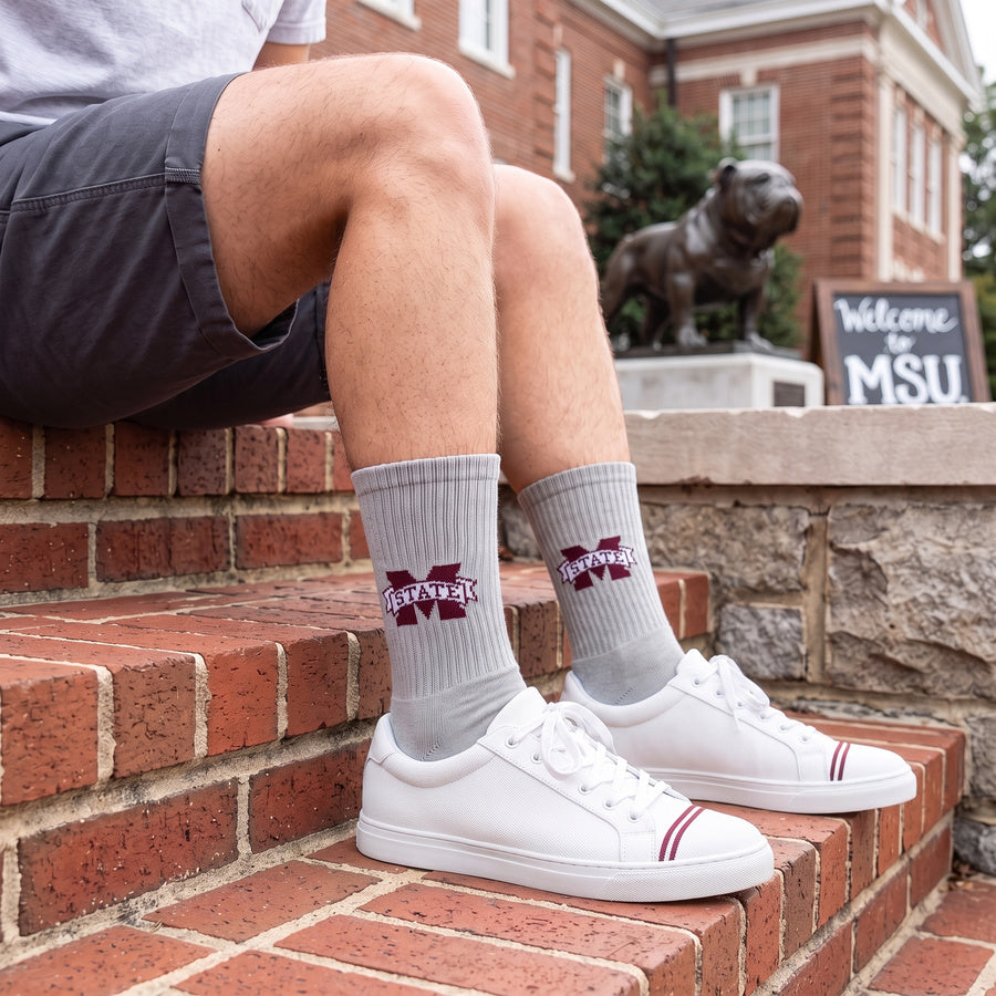 Gray M-State Logo Casual socks worn seated on campus, showing the maroon M-STATE banner logo on a heather-gray crew