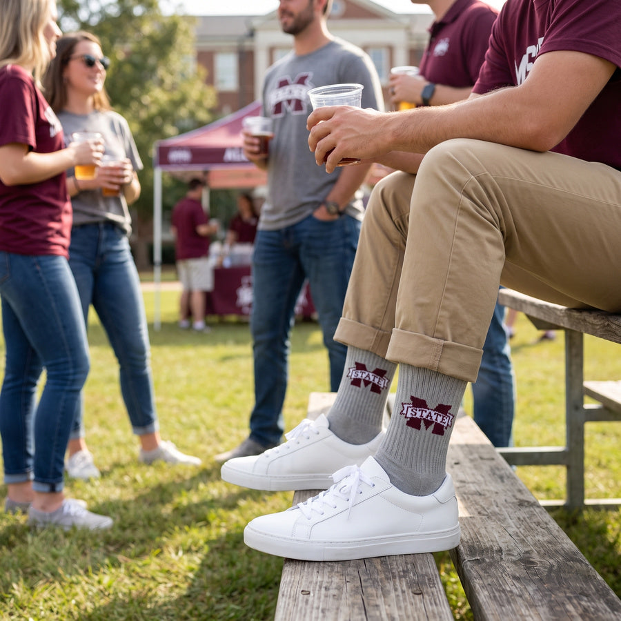 Gray M-State Logo Casual socks worn seated on campus, second angle, showing the maroon M-STATE banner logo on a heather-gray crew