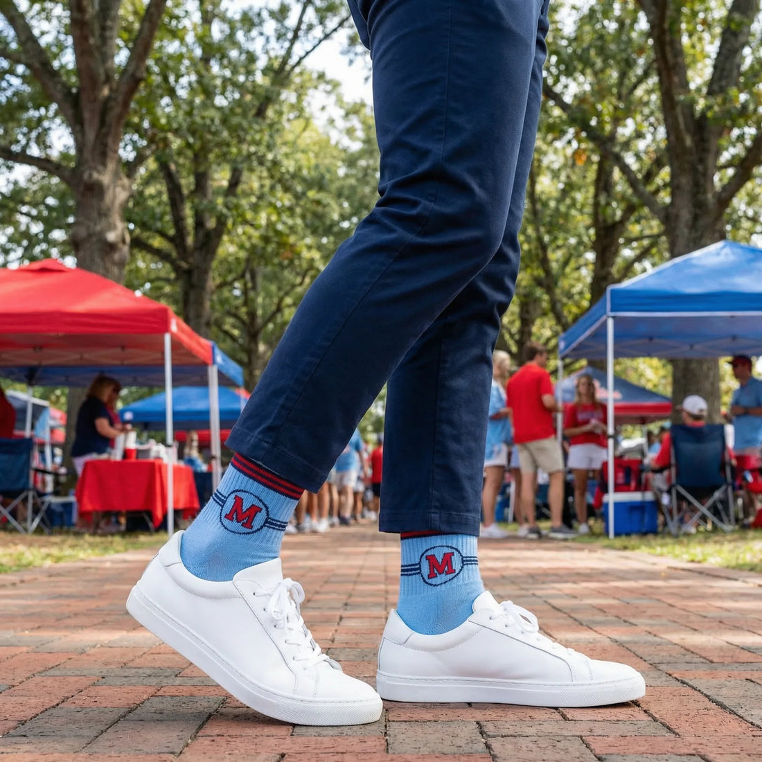 Person wearing blue socks with a logo, standing on a brick path with trees and people in the background.