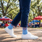 Person wearing blue socks with a red logo and white sneakers in an outdoor setting with trees and tents.