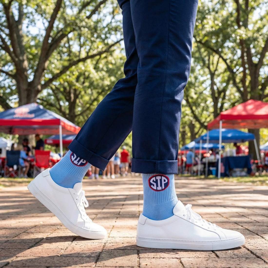 Person wearing blue socks with a red logo and white sneakers in an outdoor setting with trees and tents.