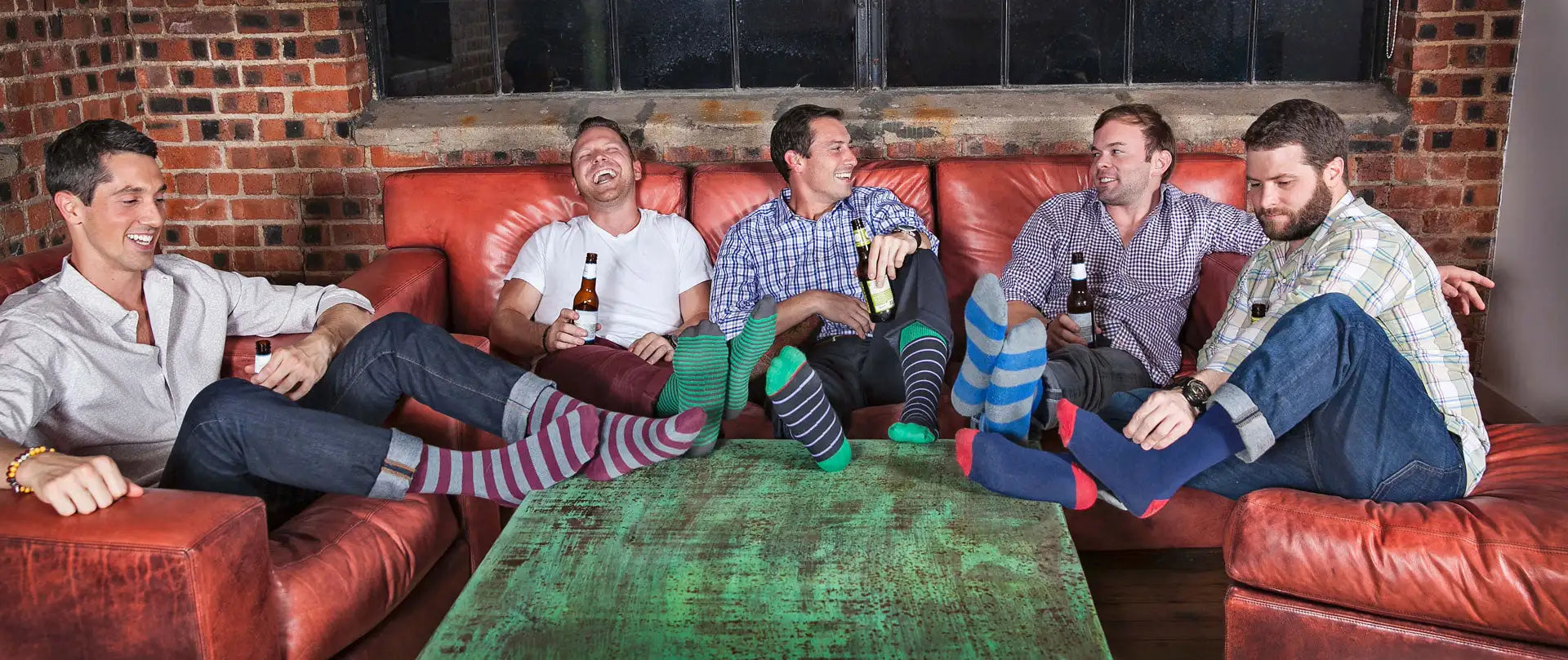 Five men lounging on a red leather couch, each wearing distinctively patterned socks—stripes and solids in green, blue, maroon, and navy—with one man’s feet bare.