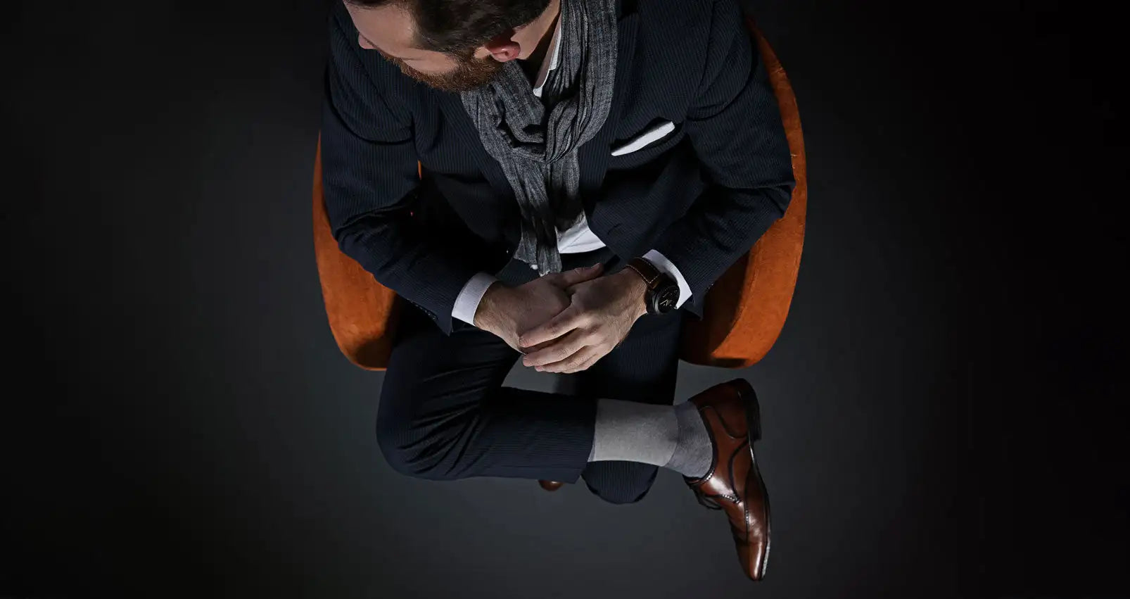 A man wearing a dark navy suit with a white pocket square and gray scarf, seated in an orange chair.