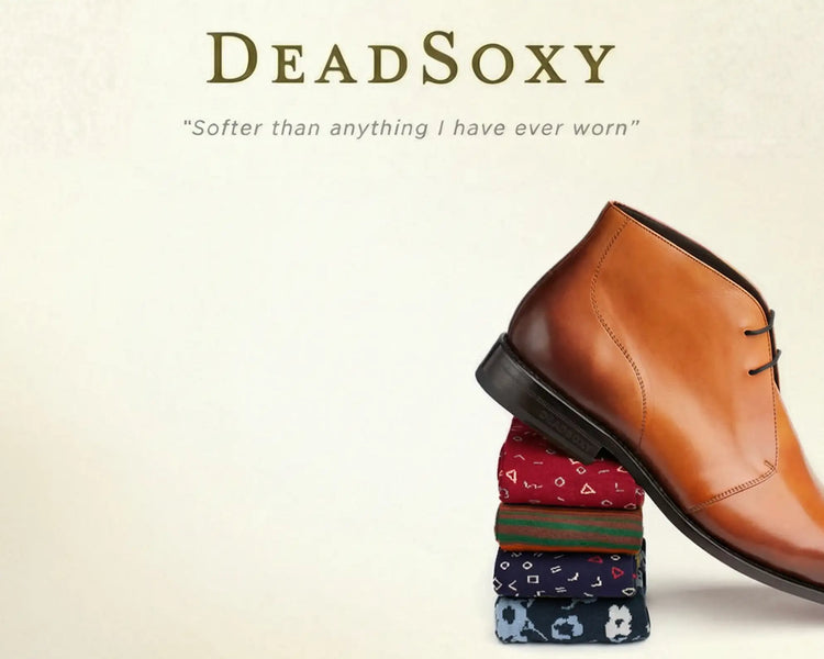 A tan leather chukka boot with black laces and a dark brown sole, positioned atop a stack of patterned socks.