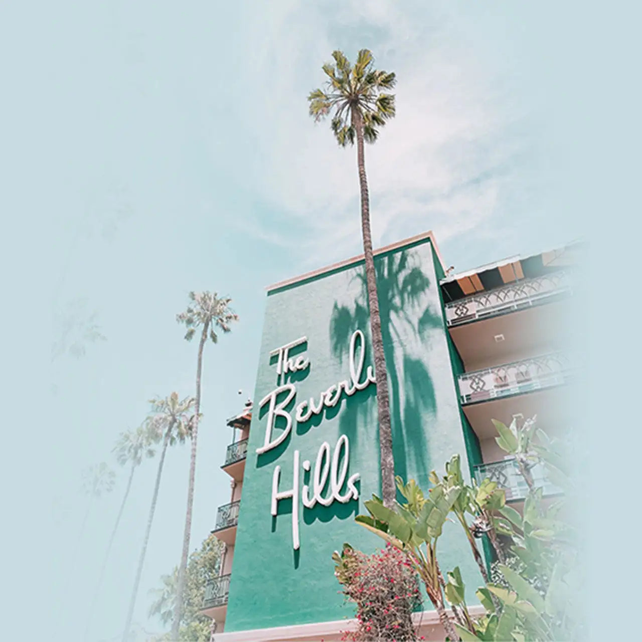 A teal-green building with white cursive lettering that reads ’the beverly hills’ and tall palm trees casting shadows on its facade.