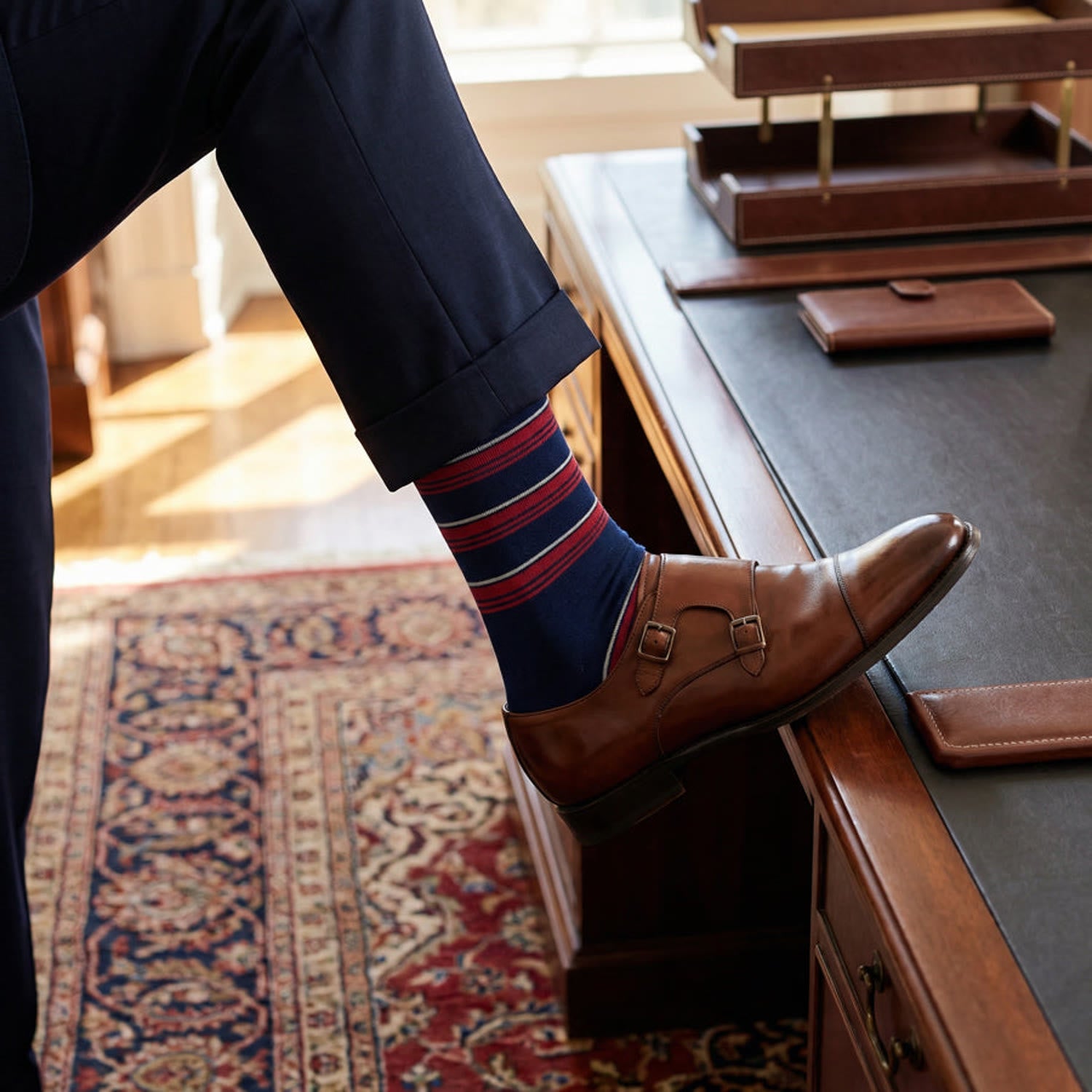 Navy dress socks with red stripes, navy trousers and brown double-monk shoes in a warm office setting